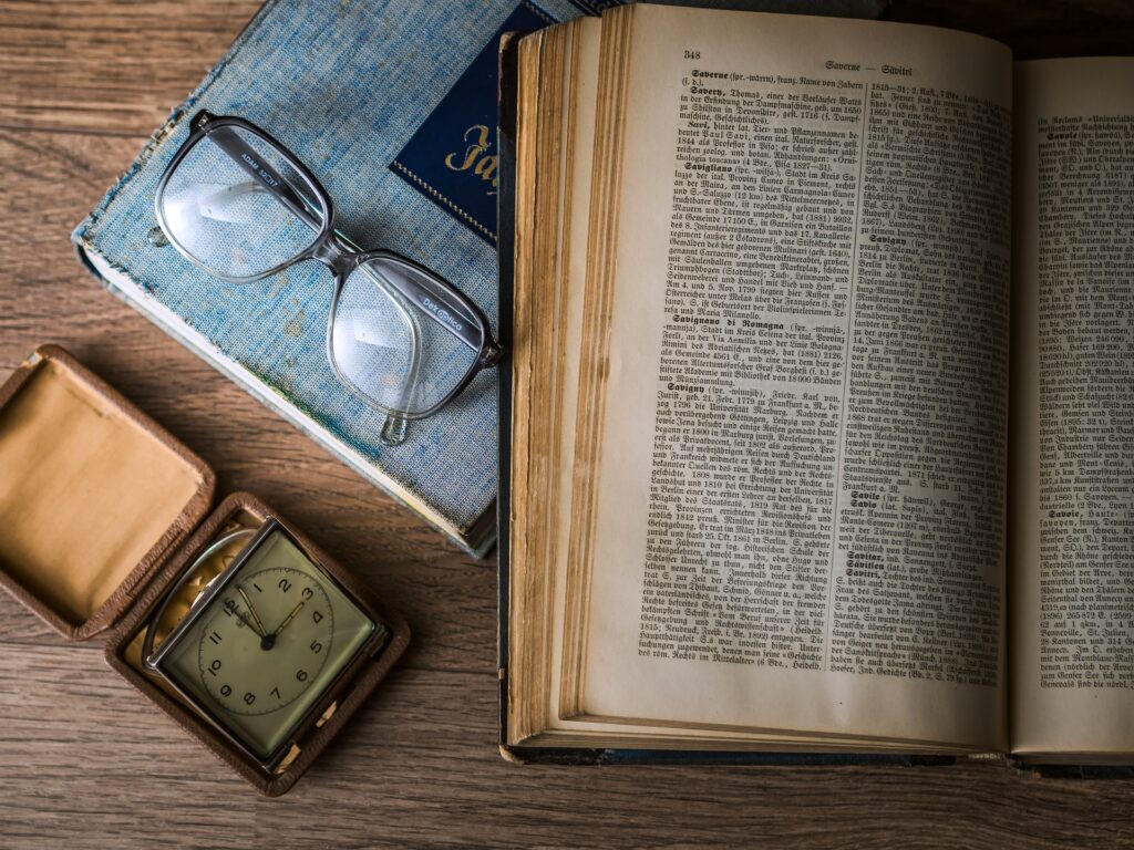 Brille, Uhr und Wörterbuch auf einem Holztisch als symbolische Darstellung von Wissen und Zeit.