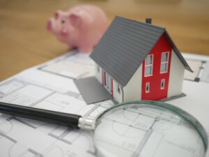 Modellhaus auf Bauplan mit Lupe und Sparschwein zur Immobilienplanung und -finanzierung.